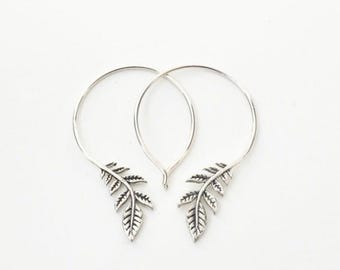 May include: A pair of silver hoop earrings with a leaf design. The earrings have a circular hoop with a detailed leaf charm at the bottom. The leaves have intricate veining and a textured appearance. The earrings are displayed against a white background.