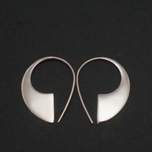May include: A pair of silver-toned hoop earrings featuring a smooth, curved, teardrop-like silhouette. The earrings have a wide, flat base that tapers into a thin, rounded wire loop, creating an open, asymmetrical shape with a polished metallic finish.