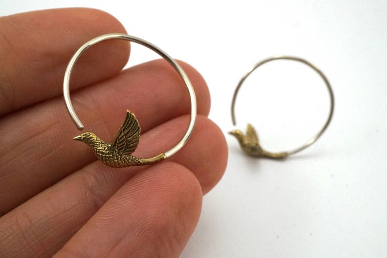 May include: A pair of silver hoop earrings with a gold bird charm attached to each hoop.