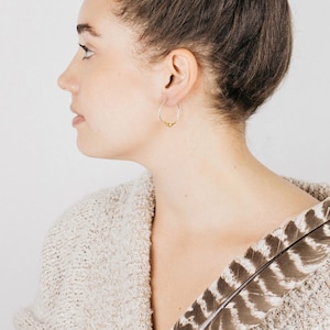 May include: A woman wearing a silver hoop earring with a small gold accent. The earring is visible in her right ear. She is wearing a cream-colored sweater with a brown and white feather pattern.