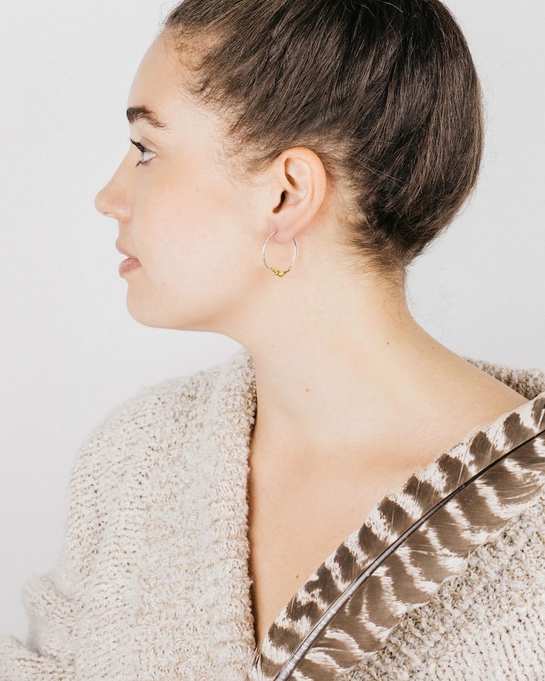 May include: A woman wearing a silver hoop earring with a small gold accent. The earring is visible in her right ear. She is wearing a cream-colored sweater with a brown and white feather pattern.