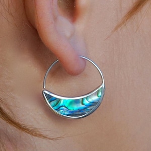 May include: A silver hoop earring featuring a crescent-shaped inlay of iridescent abalone shell. The shell shows blues, greens, and purples. The earring is worn on an ear, highlighting the jewelry's design and colour.