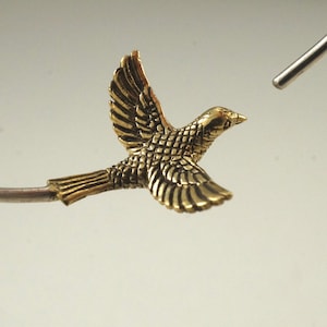 May include: A gold-colored bird charm in flight, with detailed wings and a textured body. The charm is attached to a silver-colored wire.