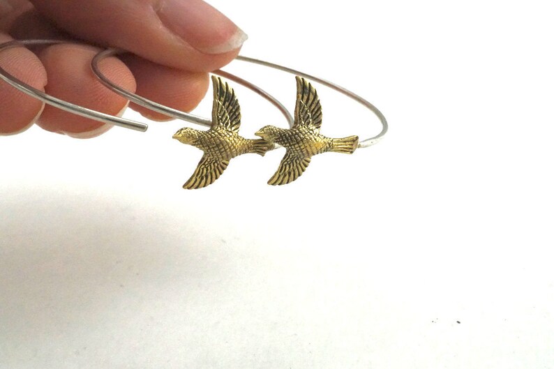 May include: A pair of silver hoop earrings with gold bird charms. The birds are facing opposite directions and have detailed wings.