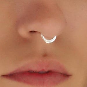 Puede incluir: Un anillo de septum en tono plateado pulido con un diseño de banda circular suave y continua. La joya está colocada en el tabique nasal, mostrando un acabado metálico reflectante y una estética sencilla para el piercing facial.