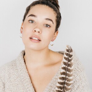 May include: A woman with brown hair wearing a beige sweater and silver hoop earrings with a gold accent. She is holding a brown and white feather.