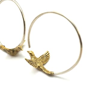 May include: A pair of silver hoop earrings with gold bird charms. The birds are facing opposite directions and have detailed wings.