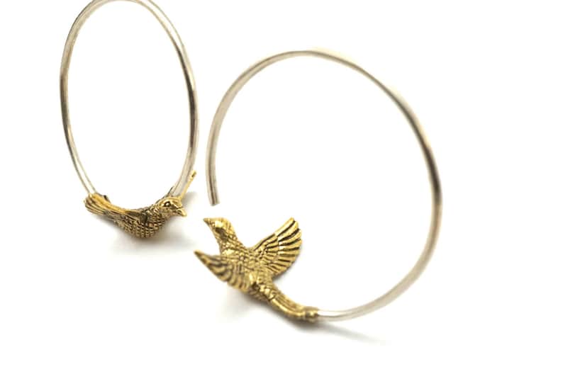 May include: A pair of silver hoop earrings with gold bird charms. The birds are facing opposite directions and have detailed wings.