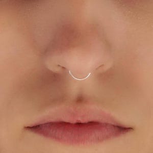 Puede incluir: Un anillo de septum circular, fino y liso, de color plateado, colocado en el centro de la nariz. La joya presenta un diseño de aro simple y continuo con un acabado metálico pulido que descansa sobre la piel del tabique nasal.