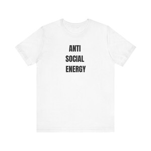 May include: A white short-sleeved t-shirt featuring the text ANTI SOCIAL ENERGY printed in bold black capital letters centered on the chest. The garment has a classic crew neckline and a standard fit, providing a clean and simple appearance.
