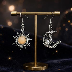 Puede incluir: Un par de pendientes desiguales de tono plateado colgados en un soporte dorado. Un pendiente presenta un diseño de sol con una piedra redonda color melocotón, mientras que el otro muestra una luna creciente con una pequeña perla blanca.