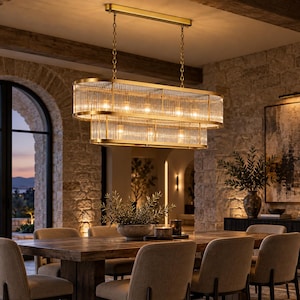 May include: An oval-shaped chandelier featuring two tiers of vertical, ribbed glass rods encased in a brushed gold metal frame. The fixture is suspended by two gold chains from a rectangular ceiling mount, emitting a warm, soft glow.