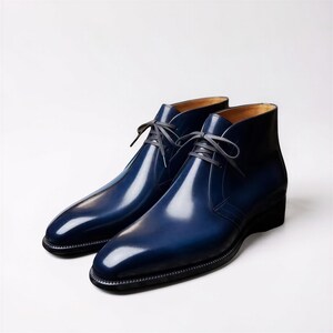 May include: A pair of polished navy blue leather chukka boots featuring a smooth, glossy finish, a rounded toe, and thin dark grey laces tied in a bow. The boots have a structured ankle-high silhouette with a dark sole and visible stitching.