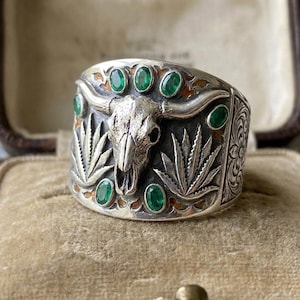 May include: A wide silver ring featuring a central steer skull motif flanked by embossed agave plants. The band is accented with six oval-cut green gemstones set around the skull and leaves, with intricate floral engravings on the side.