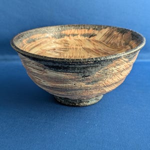May include: A ceramic bowl featuring a textured surface with a blend of black and earthy orange-brown tones. The exterior and interior display visible brushstroke patterns, a dark rim, and a raised circular base, creating a multi-layered appearance.