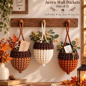 May include: A set of three crocheted acorn-shaped wall pockets hanging from a wooden rack. The pockets are brown, cream, and orange with textured caps. They contain artificial foliage, a sign reading Good Things Take Time, and a Home Sweet Home sign.