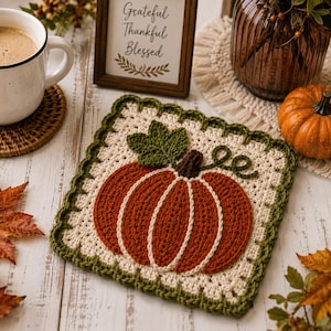 Può includere: Una presina quadrata all'uncinetto con un disegno centrale di una zucca arancione con una foglia verde scuro e un gambo marrone su sfondo color crema, rifinita con un bordo smerlato verde scuro. La zucca presenta dettagli cuciti in bianco.
