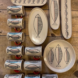 Könnte beinhalten: Ein Set aus cremefarbenem Keramikgeschirr mit schwarzen, liniengezeichneten Fischmotiven und roten Punktakzenten. Das Set besteht aus acht zylindrischen Tassen mit roten Henkeln, zwei langen schmalen rechteckigen Tabletts, einem ovalen Tablett und einem runden Teller.