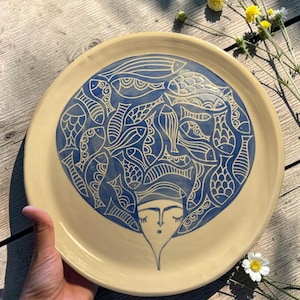 Può includere: Un piatto rotondo in ceramica con base color crema caratterizzato da un'illustrazione blu centrale. Il design raffigura un volto stilizzato con occhi chiusi e una piccola bocca, sormontato da un grande arrangiamento circolare di pesci.