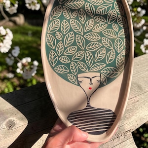 Pode incluir: Uma bandeja oval de cerâmica com base bege claro apresentando uma ilustração estilizada de uma figura. A figura tem um cabelo verde grande e arredondado preenchido com contornos de folhas brancas, olhos fechados, lábios vermelhos e camisa.