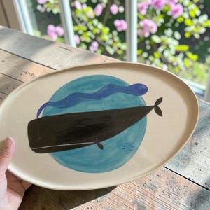 Op de afbeelding: Een ovaalvormige keramische schaal met een gebroken witte basiskleur. Het midden is voorzien van een handgeschilderde illustratie van een donkerbruine walvis die over een cirkelvormige lichtblauwe achtergrond zwemt, met een golvende blauwe lijn.