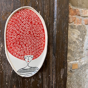May include: An oval ceramic wall plate featuring a stylized illustration of a person with voluminous red hair covered in white spiral patterns. The person has a simple facial expression with red lips and wears a black and white striped shirt.