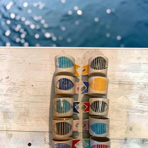 Op de afbeelding: Tien keramische bekers gerangschikt in twee kolommen, elk voorzien van een handgeschilderd visontwerp met verticale strepen. De bekers zijn gebroken wit met verschillend gekleurde vissenlichamen en contrasterende staartvinnen in blauw, geel, rood, roze en zwart.