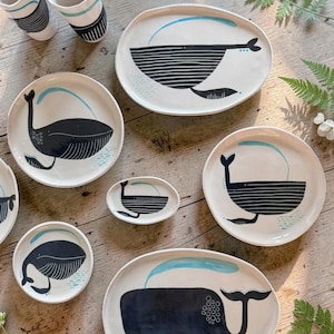 Puede incluir: Un conjunto de vajilla de cerámica que incluye dos vasos altos, dos fuentes ovaladas grandes y tres platos redondos de varios tamaños, todos con ilustraciones de ballenas en blanco y negro con detalles en azul claro sobre un fondo color crema.