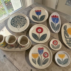 Op de afbeelding: Een set crèmekleurig keramieken servies met handgeschilderde bloem- en plantmotieven in rood, blauw en geel. De set bevat ronde borden, ovale schalen, kleine ronde schoteltjes en drie cilindervormige bekers op een bijpassend dienblad.