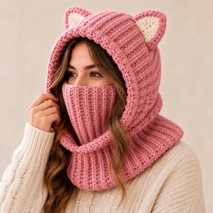 Peut inclure: Une cagoule en crochet rose vieux rose avec une texture en tricot côtelé et deux oreilles de chat sur le dessus. Les oreilles ont des centres de couleur crème. La capuche couvre la tête et le cou, avec une section masque intégrée pour le nez.