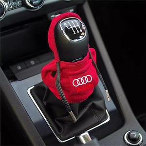 Puede incluir: Una funda de tela roja para la palanca de cambios con forma de sudadera con capucha, que presenta el logotipo y el texto de Audi en blanco en el bolsillo frontal. Incluye cordones negros con puntas metálicas y un patrón de seis velocidades.