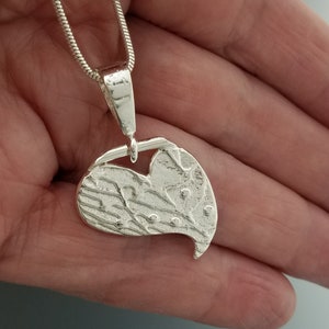 May include: A silver pendant necklace featuring a textured heart-shaped charm. The charm has a unique, organic design with a textured surface and small, raised dots. The pendant hangs from a delicate silver chain.