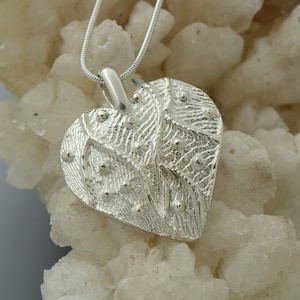May include: A silver heart-shaped pendant with a textured surface and small, raised dots. The pendant hangs from a thin, silver chain. The heart design features a detailed, organic pattern, creating a unique and artistic piece of jewelry.