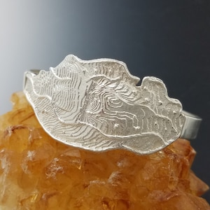 May include: A silver-colored cuff bracelet with a detailed, textured design resembling a topographical map. The bracelet is set against a backdrop of a golden-yellow, crystalline surface.