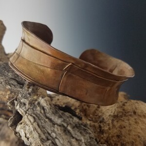 May include: A copper-colored cuff bracelet with a textured, layered design. The bracelet has a wide band with a curved shape and is displayed on a piece of driftwood. The metal has a warm, antique finish.