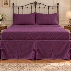May include: A deep purple bed set featuring a fitted sheet, two matching pillowcases, and a bed skirt with decorative side ties. The bedding is displayed on a metal frame bed, showcasing a solid colour fabric with a soft, textured appearance.