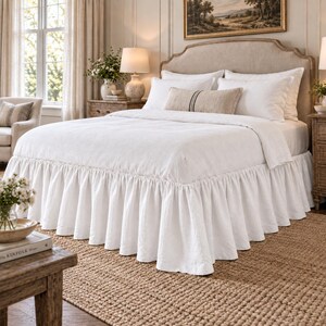 May include: A white bed valance with a gathered ruffle design, featuring a long, flowing drop that reaches the floor. The bed valance is made of a solid white fabric and is positioned beneath a white duvet cover on a bed frame with a beige headboard.