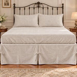 May include: A bed featuring a light beige bed skirt with decorative side ties, paired with a matching flat sheet and two pillows. The bed is framed by a dark metal headboard, positioned between two wooden side tables with lamps and flowers.