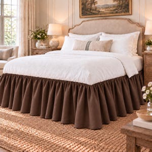 May include: A bed featuring a solid brown bed skirt with a gathered ruffle design. The bed is dressed in white bedding, including a white duvet cover and white pillows, with one rectangular accent pillow featuring a thin dark vertical stripe.