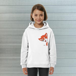 Könnte beinhalten: Ein weißer Kapuzenpullover mit einer Grafik eines orangefarbenen Fuchses, der seitlich hervorlugt. Der Fuchs hat weiße Akzente und eine schwarze Nase. Unter dem Fuchs ist das Wort TAPS in kleinen schwarzen Buchstaben auf der Vorderseite gedruckt.