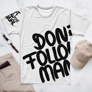 Könnte beinhalten: Ein weißes Kurzarm-T-Shirt mit großen schwarzen Pinselstrich-Buchstaben, die "DON'T FOLLOW MAN" auf der Vorderseite bilden, sowie dem Text "READ THE MANUAL" in Schwarz auf dem linken Ärmel, präsentiert auf einer flachen Oberfläche.