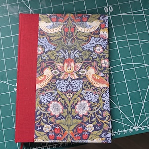 May include: A hardback book featuring a red fabric spine and a front cover with a repeating pattern of birds, strawberries, and floral motifs in shades of orange, green, and light blue against a dark purple background.