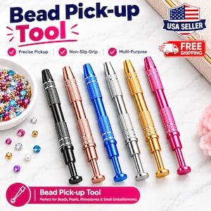 May include: Six metal bead pick-up tools in black, rose gold, blue, silver, gold, and pink. Each tool features a textured grip and a plunger mechanism. A white bowl filled with colorful rhinestones and pearls sits to the left of the tools.