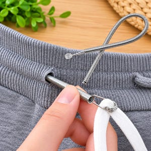May include: A flexible metal spring drawstring threader tool with a looped end, shown being used to guide a white flat cord through the ribbed waistband of gray fabric sweatpants. The tool features a coiled wire design for threading.