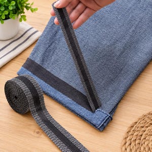 May include: A roll of black elastic band with a textured white woven pattern along the edges. The elastic is shown being applied to the hem of a blue denim fabric garment, demonstrating its flexibility and use for clothing construction.