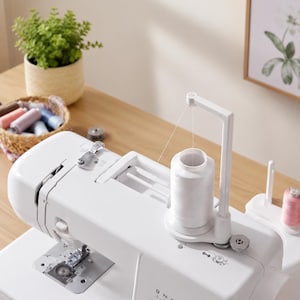 May include: A white sewing machine featuring a vertical spool pin holding a large white thread spool. The machine includes a metal needle plate, a thread guide arm, and a bobbin winding mechanism. A basket of colorful thread spools sits nearby.