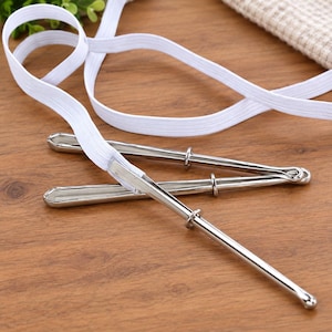 May include: A set of three silver-toned metal bodkins with smooth, rounded tips and a small sliding ring on each shaft. A long, white, flat elastic band is threaded through the eye of one of the metal bodkins, connecting the tools together.