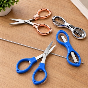 May include: A collection of three pairs of scissors featuring metallic rose gold, polished silver, and bright blue handles. Each pair has sharp stainless steel blades and symmetrical finger loops, arranged on a flat surface with a thin metal rod.