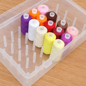 May include: A clear plastic storage box containing twelve spools of sewing thread in various colors including purple, white, orange, yellow, red, brown, and pink. The spools are arranged on vertical pegs inside the rectangular container.