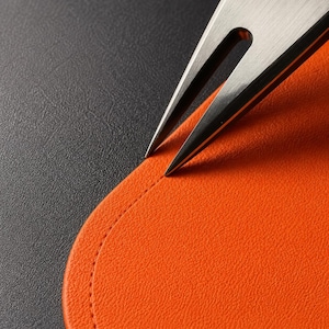 May include: A silver-toned metal fork with two sharp tines resting against the curved edge of a bright orange textured material. The orange piece features a visible stitched seam running parallel to its rounded border on a dark surface.